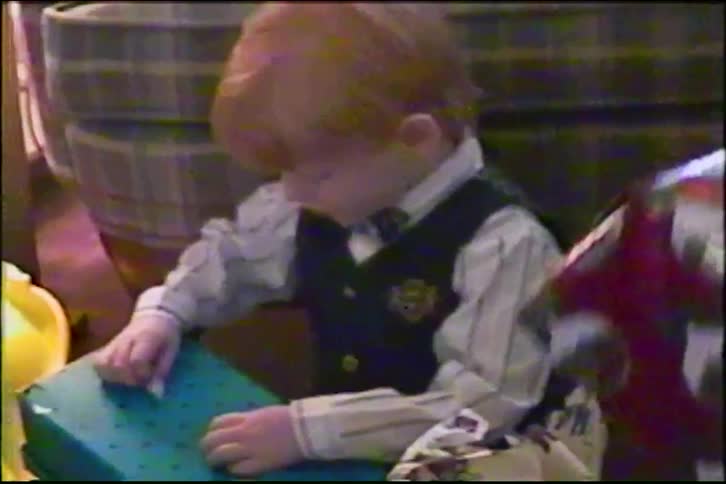 Vintage VHS footage shows a cute red-haired boy in a vest and bow tie excitedly opening a Christmas present