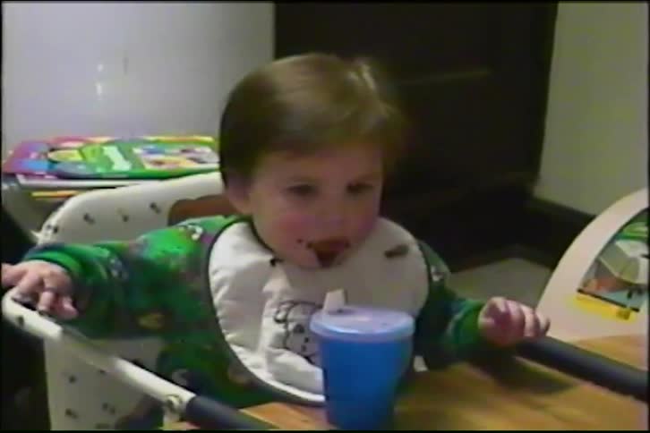 A cute toddler with a messy face eats from a spoon in a high chair in a vintage home video