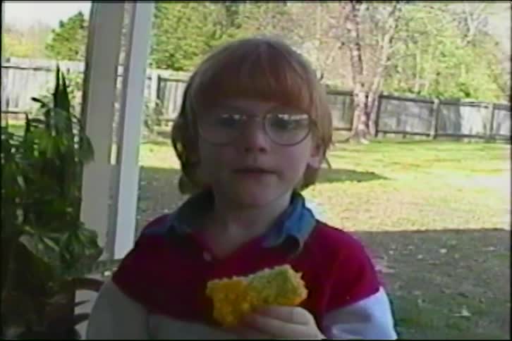 A young boy with red hair and glasses eats a colorful snack outside while a girl and dog play in the backyard