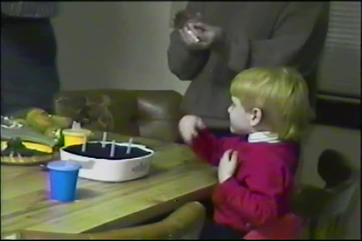 A cute young boy with blond hair excitedly blows out the four candles on his birthday cake in a vintage home movie