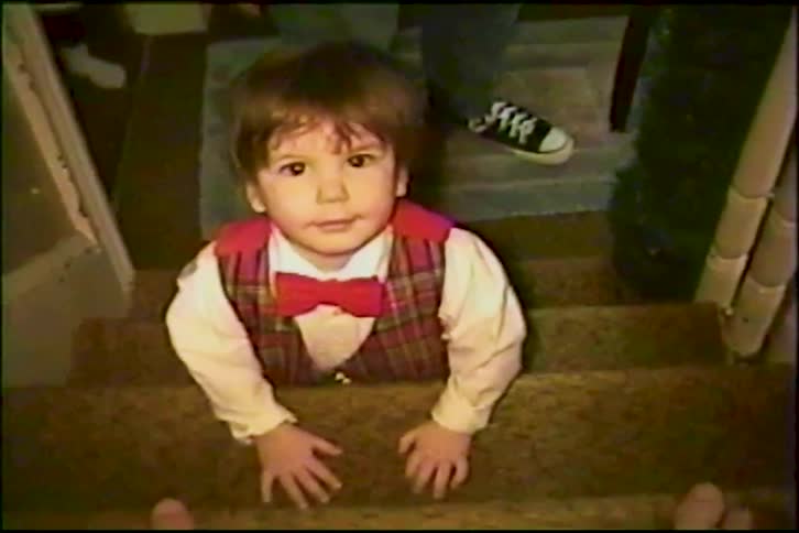 A vintage VHS home video shows a cute toddler in a plaid vest and bow tie crawling up the stairs before being picked up by their dad