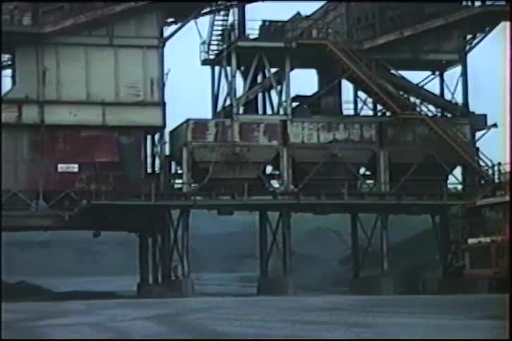 Vintage footage pans from a large water truck to a massive, rusty industrial plant at a desolate quarry