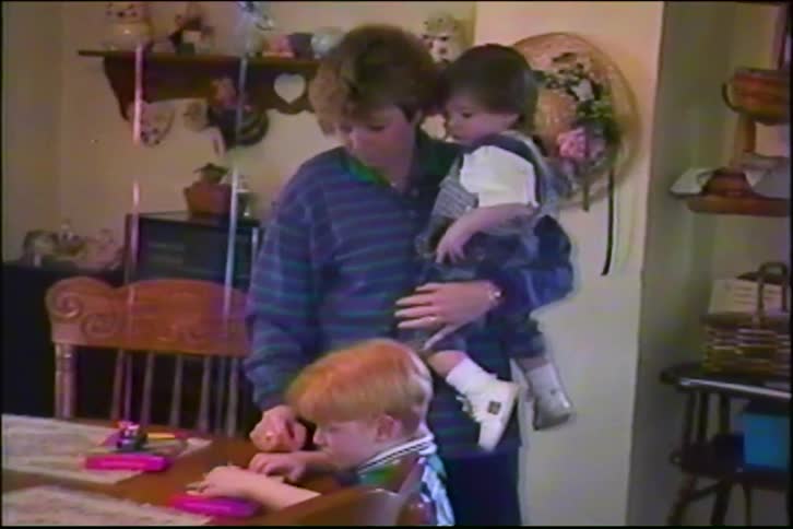 A mother holds her toddler daughter as her young son sits at the dining table to play with art supplies in this sweet vintage home video from the 1990s