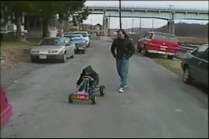 A father watches his young child learn to use a pedal go-kart on a quiet neighborhood street in a nostalgic home video