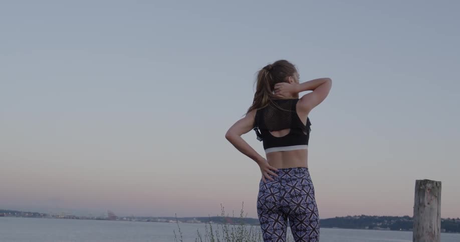 A woman stands near the water holding her neck