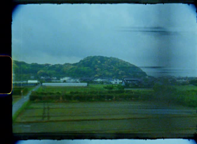 A vintage 8mm film captures the view of a lush green Japanese countryside with a distant hill, seen from the window of a moving train