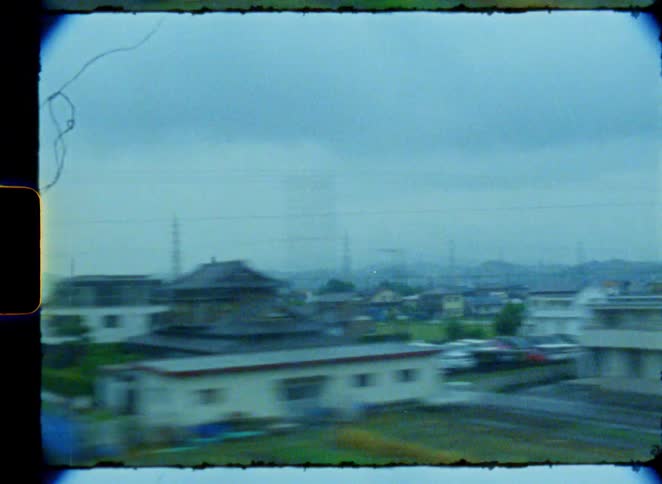 A vintage 8mm film view from a fast-moving train captures the blurry, passing scenery of a Japanese town on a gloomy day