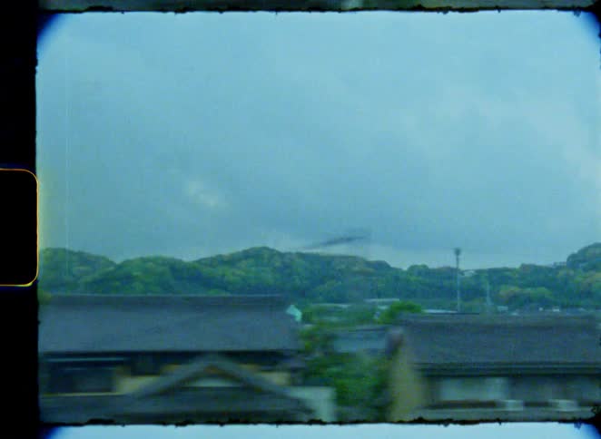 A grainy, vintage film captures the view of a Japanese town and lush green hills from the window of a moving train