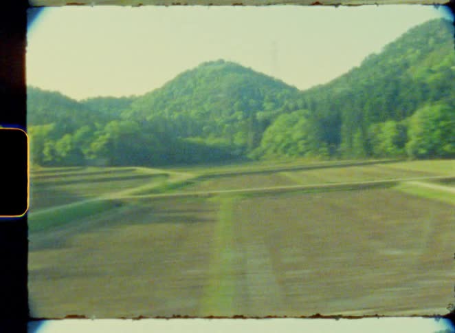 A grainy, nostalgic view from a moving vehicle captures the Japanese landscape, from rural rice fields to suburban roads, on vintage 8mm film