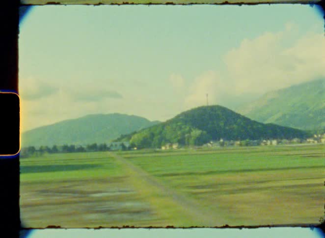Vintage 8mm film captures the scenic view of a rural, mountainous landscape from the window of a moving vehicle