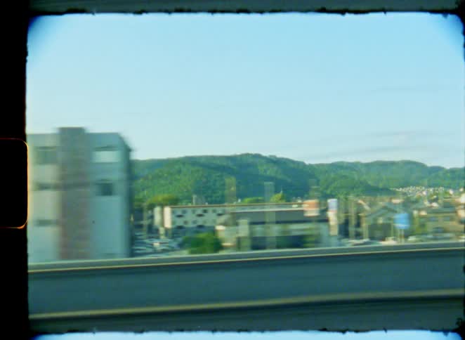 A vintage film view from a moving train shows a sun-flared city skyline and the Japanese suburban landscape passing by