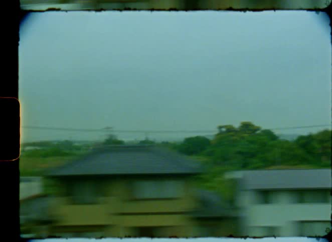 A vintage film view from a moving train window captures the passing rural and industrial landscape of Japan on a cloudy day