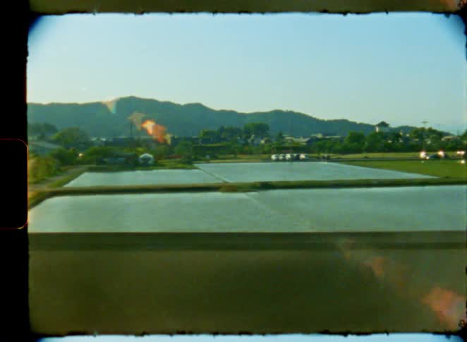 A nostalgic journey through the serene Japanese countryside is captured on vintage 8mm film from a train window, revealing views of rice paddies and distant mountains
