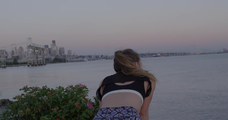 A woman in a sports bra leans over to catch her breath while overlooking a river at sunset