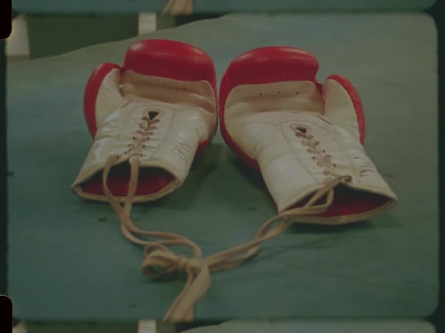 A pair of vintage red and white boxing gloves with laces sit on the green canvas of a boxing ring