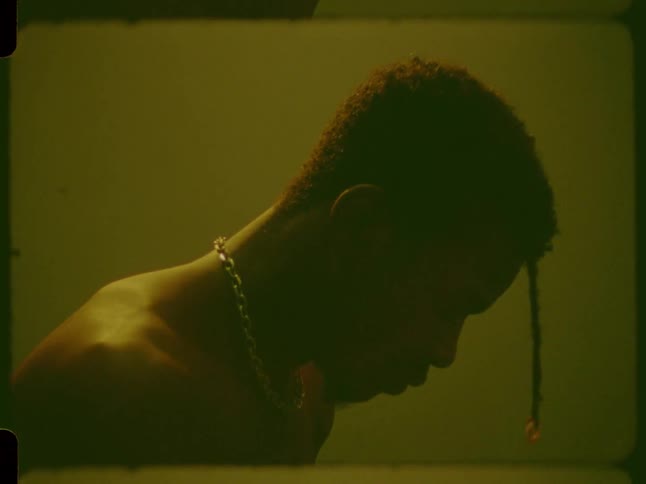 Moody 16mm film shot of a contemplative young man with a chain necklace lowering his head in a shadowy, green-lit room