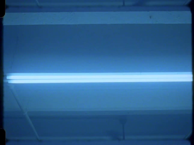A vintage film shot looks up at a ceiling fan rotating under a bright, blue fluorescent light