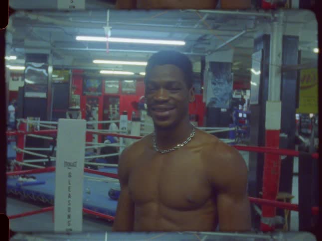 A young, muscular Black boxer smiles confidently for the camera while standing in a gritty, retro-style boxing gym