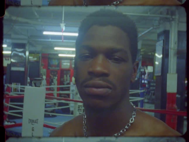 A determined male boxer with a muscular physique stares into the camera as it zooms in for a close-up portrait inside a gritty boxing gym