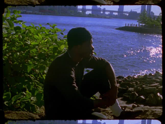 Shot on 16mm film, a young man sits thoughtfully on a rocky riverbank in a city park on a sunny day