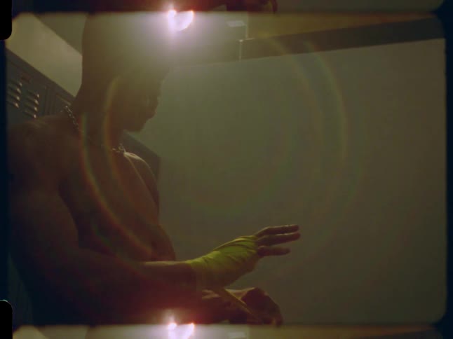 Vintage-style 8mm footage shows a muscular fighter wrapping his hands in a moody, backlit locker room
