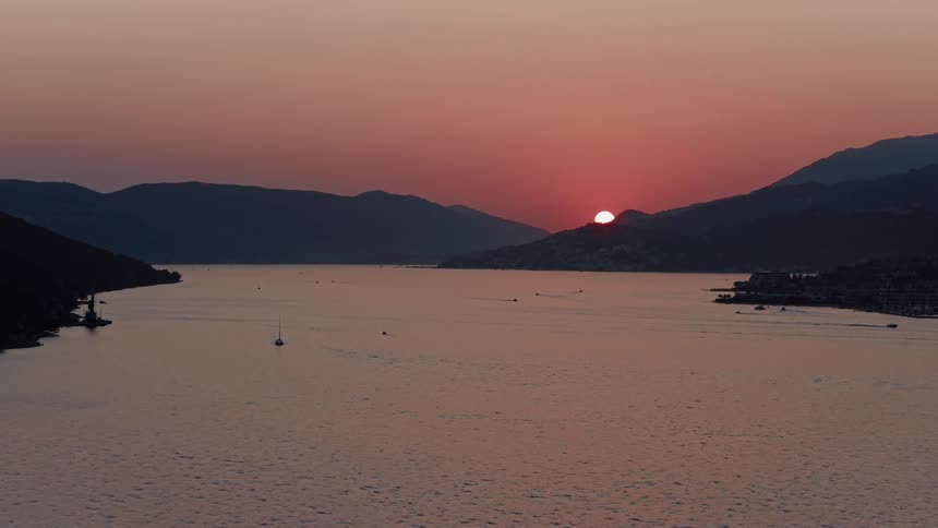 A serene time-lapse captures the sun setting behind silhouetted mountains over a calm bay with boats