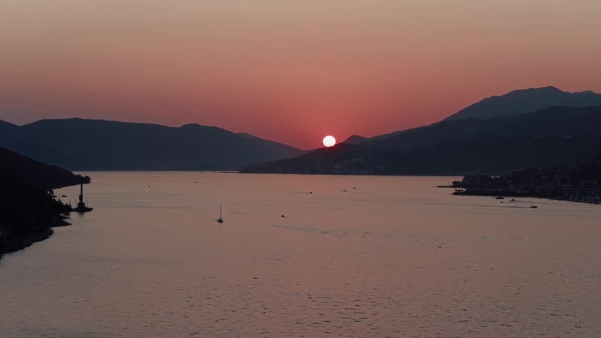 An aerial shot captures the serene sunset as the sun dips behind the mountains over a tranquil bay with boats on the water