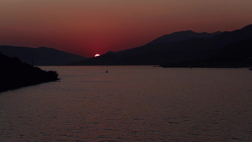 The last sliver of a deep red sun dips behind a mountain silhouette over a tranquil bay