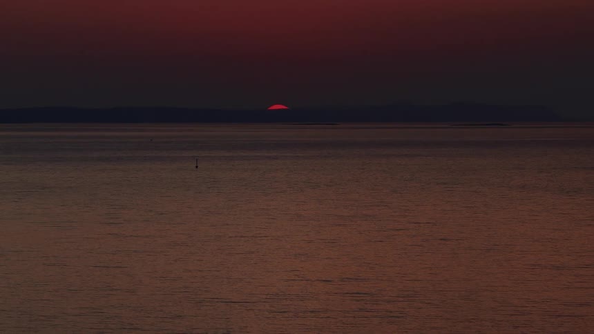 A slow zoom captures the last sliver of a deep red sun as it disappears behind the distant horizon over the tranquil ocean at dusk