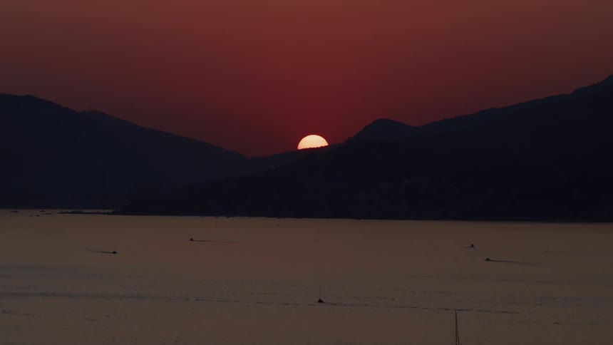 A beautiful aerial view captures the sun slowly setting behind silhouetted mountains, casting a deep red glow over a tranquil bay