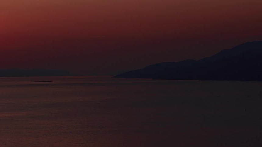 The calm ocean reflects the deep crimson sky at dusk, with dark mountain silhouettes on the horizon