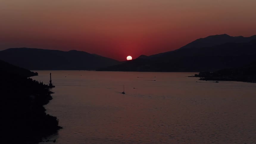 The sun sinks behind silhouetted mountains, casting a deep red glow over a tranquil bay as a lone sailboat passes