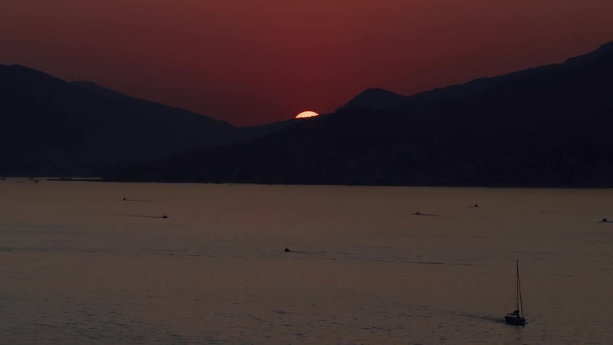 A dramatic red sun sets behind silhouetted mountains, casting a serene glow over the sea and sailboats