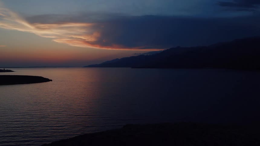 Drone slowly pulls back over a calm sea at twilight, revealing a dramatic sunset sky and silhouetted mountains on the horizon