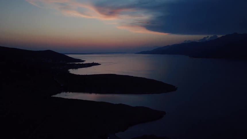 Aerial view of a tranquil bay at dusk, where the silhouetted coastline frames the calm water reflecting the colorful twilight sky