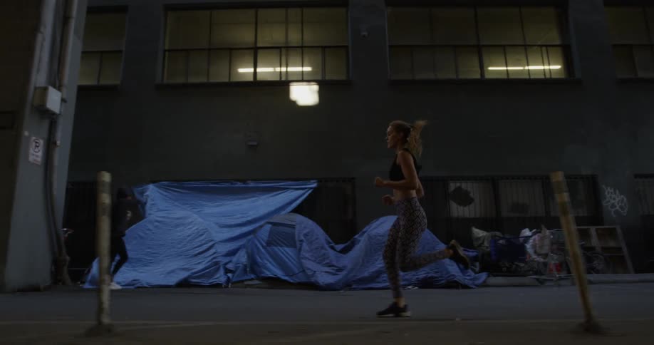 A female is jogging past homeless tents near warehouses