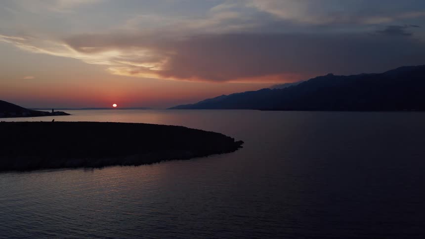 Aerial footage ascends over a tranquil sea during a stunning sunset, revealing a silhouetted coastline and distant mountains