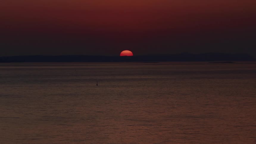 The vibrant red sun sets behind distant mountains, casting a dark, serene glow over the calm ocean water