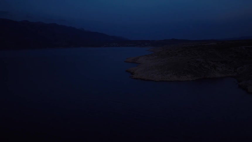 A still wide shot of a dark, rocky coastline and tranquil sea under a deep blue sky at night