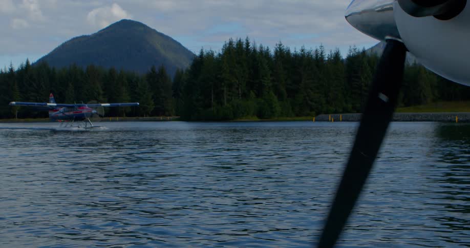 A seaplane's propeller spins in the foreground as another seaplane taxis on a tranquil mountain lake