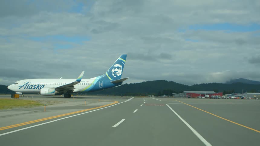 A point-of-view shot driving on an airport tarmac towards a parked Alaska Airlines jet with mountains in the background