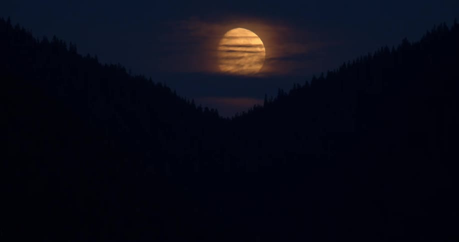 A massive, orange full moon glows behind thin clouds, perfectly framed by the silhouette of a mountain valley at night
