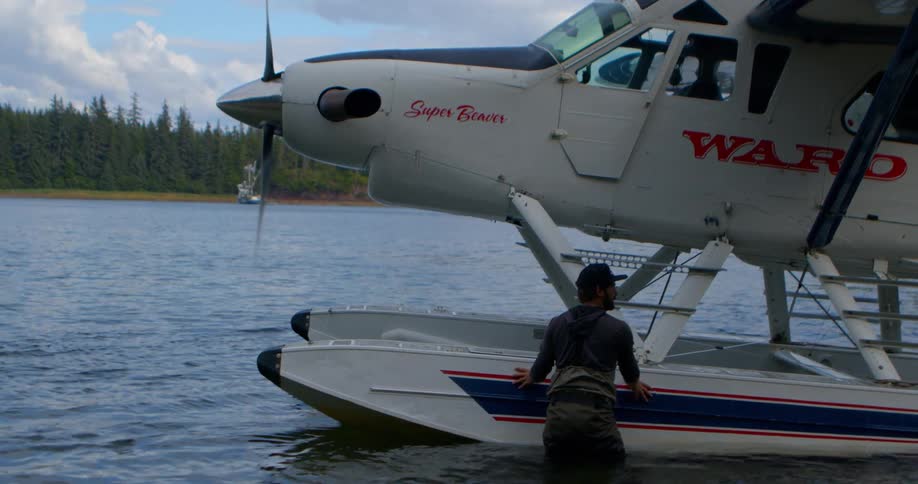 A man in fishing waders carefully guides a seaplane with its propeller spinning away from the shore of a remote lake