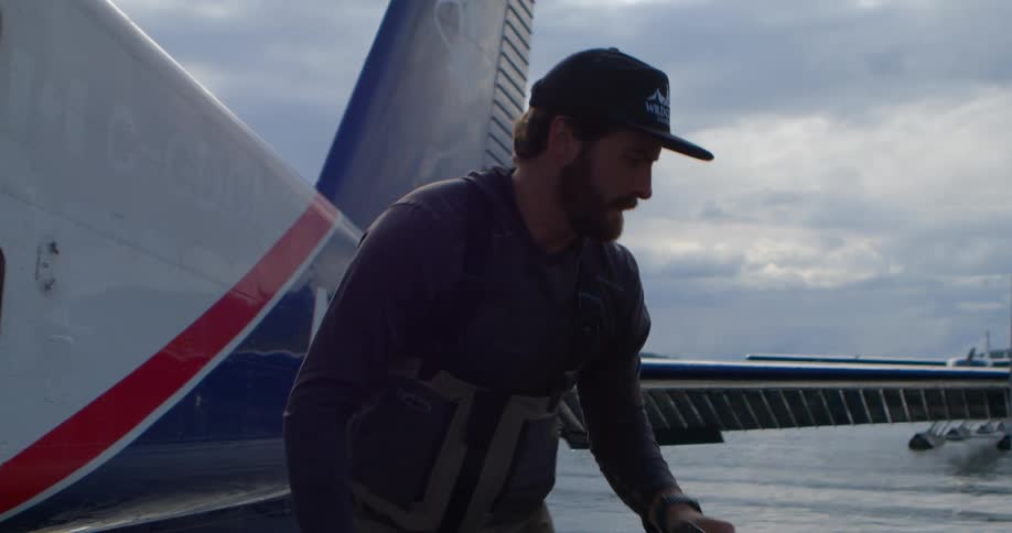 A man in fishing waders loads his gear into the side of a seaplane floating on the water in slow motion
