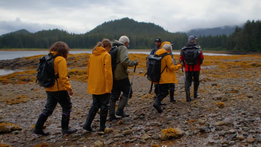 A tour group of hikers in rain gear and backpacks walk along a wet, rocky coast with forested hills in the background