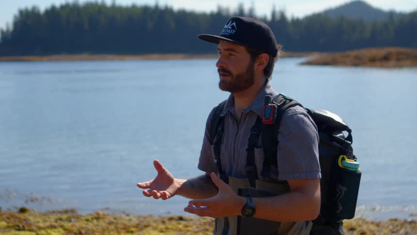 A bearded outdoorsman in waders and a backpack gestures while talking on the shore of a remote, forested inlet on a sunny day