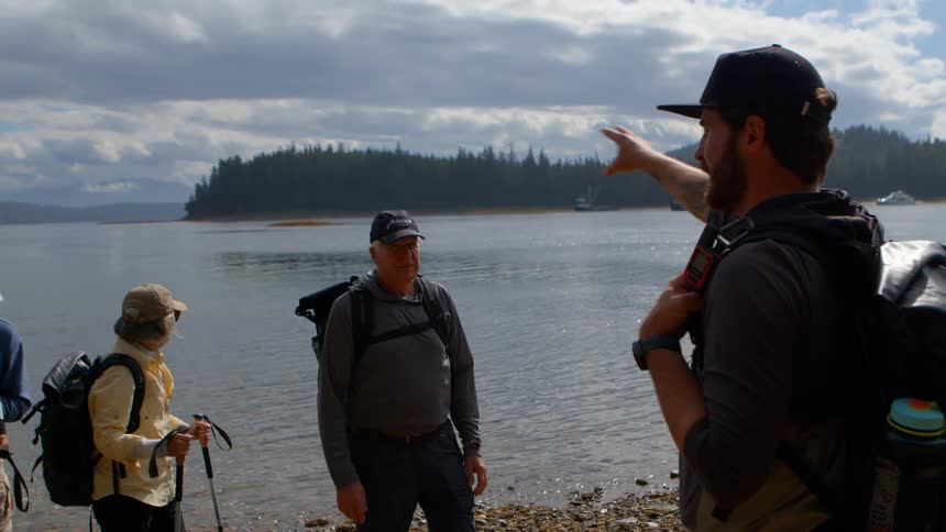 A hiking guide points across the water while talking to a group of senior hikers on a rocky shoreline