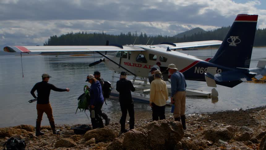 A group of adventurers unloads their gear from a seaplane onto a rocky shore in a remote wilderness location