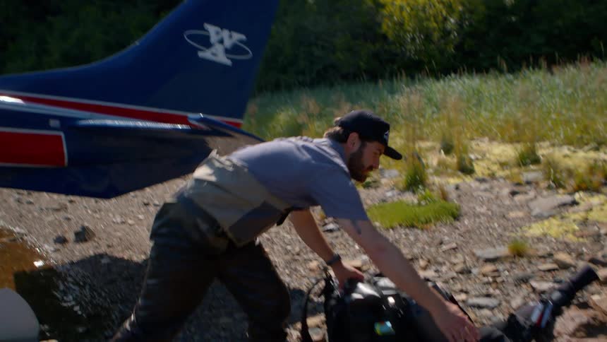A man in waders unloads expedition gear from a seaplane parked on a remote, rocky shore in slow motion
