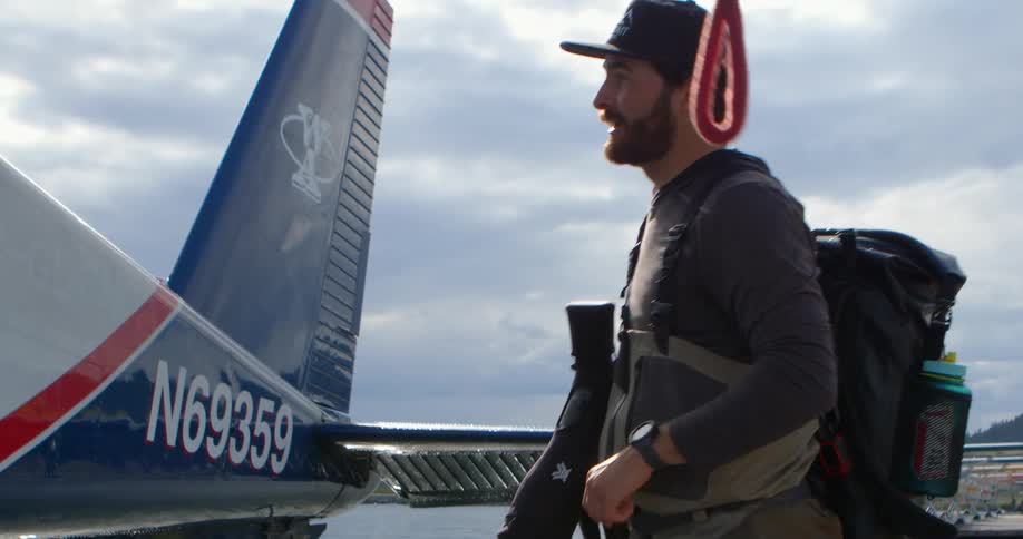 An outdoorsman in waders loads his gear and prepares to board a seaplane on a dock for a remote adventure